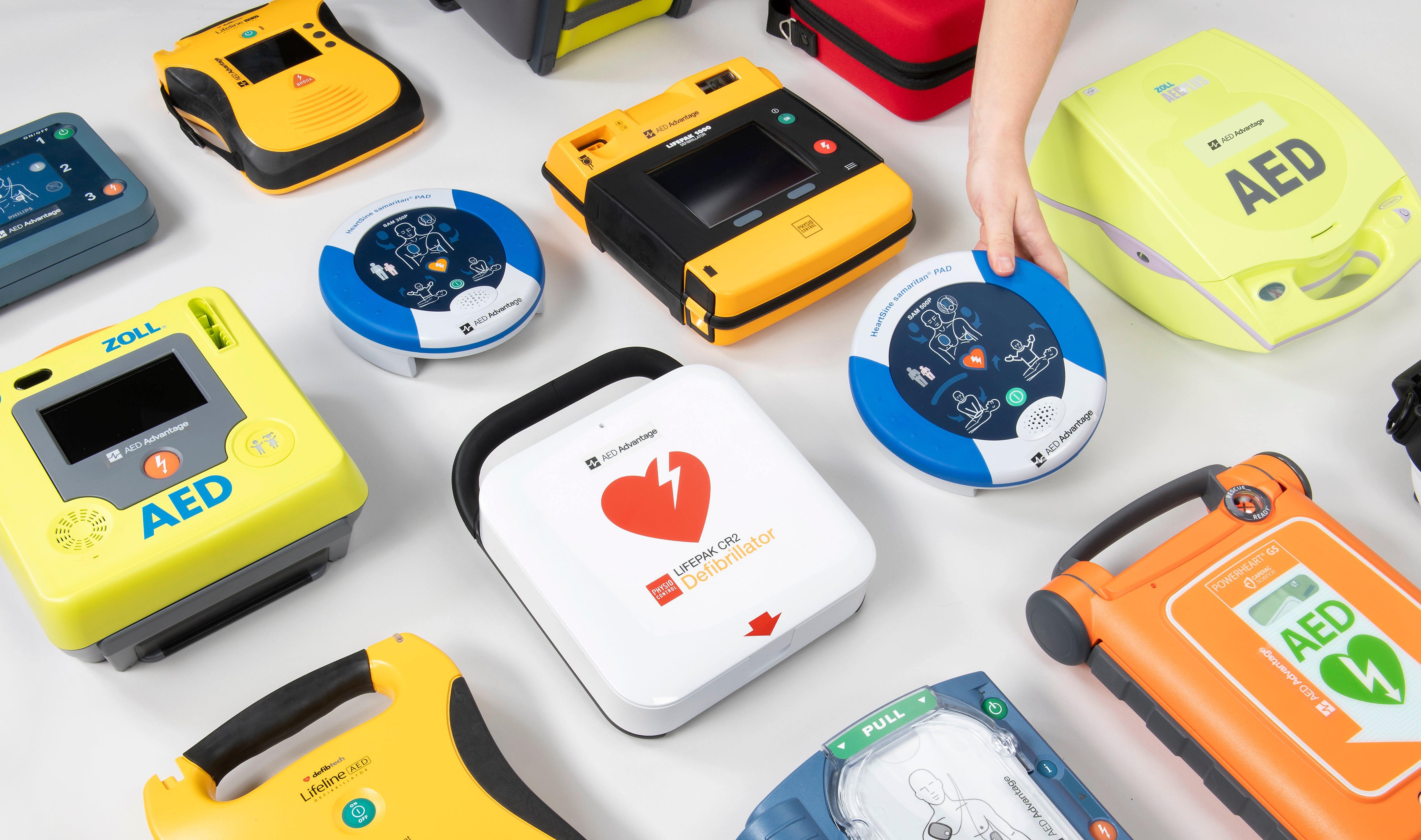A collage of AEDs of various colours. A blue and white HeartSine AED is being retrieved from the bunch. 